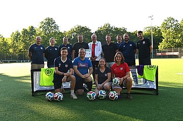 Das Foto zeigt eine Gruppe von Vereins- oder Teammitgliedern auf einem Fußballfeld. Die Gruppe ist symmetrisch vor zwei kleinen Trainings- oder Minitoren angeordnet. Zwischen und vor den Toren liegen insgesamt mehrere Fußbälle. Eine Person in der Mitte der hinteren Reihe hebt ein Dokument mit sichtbarer Auszeichnung oder Urkunde in die Kamera. Die Kleidung der Gruppe wirkt einheitlich sportlich und deutet auf einen gemeinsamen Vereins- oder Mannschaftskontext hin. Der Hintergrund besteht aus einer grünen Baumreihe und Teilen der Sportanlage. Die Atmosphäre erinnert an eine offizielle Übergabe oder Ehrung im Vereinsumfeld.