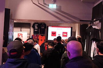 Gruppe von etwa 20 Personen in einem Laden mit Fußballartikeln (T-Shirts, Jacken) versammelt sich vor Bildschirm mit „ALBANIEN vs.“-Anzeige. Fokus auf Rückenansichten und Profile, beleuchteter Innenraum mit Ausgangshinweisen oben. Ideal für Event-Promo im Berliner Fußball-Verband-Kontext.