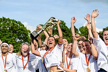 Group on soccer field lifting trophy, wearing medals; white jerseys, arms raised, trees and blue sky in background.