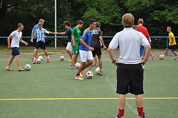 Auf einem Fußballplatz trainiert eine Gruppe Jugendlicher. Mehrere Jugendliche bewegen sich mit Bällen über das Kunstrasenfeld und führen einfache Ballübungen aus. Im Vordergrund steht eine Person in Sportkleidung mit den Händen in die Hüften gestützt und beobachtet die Trainingsgruppe. Im Hintergrund ist eine Baumreihe zu sehen, die den Platz umgibt.