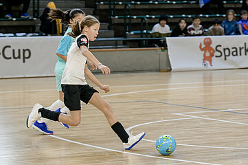 The picture shows a scene in a sports hall during an indoor soccer match. In the foreground, two people are actively involved in the game. One person is wearing a white jersey with black sleeves and black shorts as well as white sports shoes with blue soles. This person is in motion and appears to be playing or dribbling the ball. The ball is blue with green and yellow patterns. Right next to them is another person in a turquoise jersey with matching shorts and white sports shoes. The floor is made of light-colored wood, typical of indoor sports fields, and the field lines are marked in white. Empty green stands can be seen in the background with a few people sitting on them.