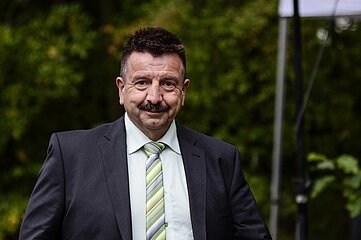 Auf dem Foto steht eine einzelne Person im Vordergrund, deren Oberkörper sichtbar ist. Die Person trägt einen formellen Anzug in dunkler Farbe sowie ein helles Hemd. Dazu passend ist eine gestreifte Krawatte in verschiedenen Grün‑ und Grautönen zu sehen. Der Hintergrund besteht aus dichter, grün wirkender Vegetation, vermutlich Bäume oder Sträucher. Rechts im Bild ragt ein vertikales technisches Element ins Bild, möglicherweise ein Stativ oder Teil einer Konstruktion. Die Beleuchtung wirkt natürlich, als wäre die Szene im Freien aufgenommen worden. Die Person steht aufrecht und ist zentral im Bild platziert. Weitere Gegenstände oder Personen sind nicht sichtbar.