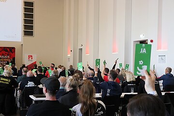 "Eine Gruppe von Menschen sitzt an Tischen im Rathaus Schöneberg und hält grüne Karten mit der Aufschrift 'JA' hoch. Im Hintergrund sind weiße Wände und ein Bildschirm mit Text zu sehen, auf welchem die Powerpoint-Präsentation, die durch den Abend begleitet, läuft.