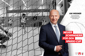In einer Sporthalle ist im Hintergrund ein laufendes Hallenfußballspiel zu sehen. Mehrere spielende Jugendliche bewegen sich dynamisch vor einem großen Handball- oder Fußballtor. Im Vordergrund steht eine Person im dunklen Anzug und weißem Hemd, mit verschränkten Händen, vor dem Netz des Tors. Rechts im Bild befindet sich eine helle grafische Fläche mit eingebettetem Stadtplanmotiv. Darauf steht der gedruckte Text: „Kai Wegner, Regierender Bürgermeister von Berlin“ sowie in einem roten Schriftfeld: „Neuer Schirmherr für den Sparki‑Cup“. Unten rechts sind die Logos mehrerer Institutionen abgebildet.