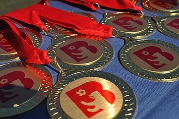 Auf dem Foto liegen mehrere goldfarbene Medaillen auf einem blauen Stoff. Jede Medaille hat ein rotes Band, das oben sichtbar zusammengelegt ist. Auf den runden Medaillen befindet sich in der Mitte das Logo des Berliner Fußball-Verbandes. Die Medaillen liegen dicht nebeneinander und reflektieren das Sonnenlicht, wodurch einzelne Bereiche glänzend wirken. Der Hintergrund ist unscharf, sodass der Fokus klar auf den Medaillen liegt.