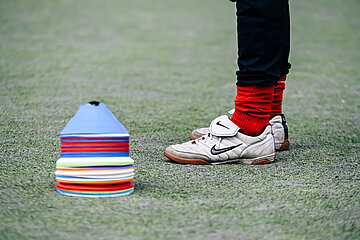 Auf dem Foto ist der untere Teil einer Person zu sehen, die auf einem Sportplatz mit Kunstrasen steht. Sichtbar sind die Beine der Person, bekleidet mit schwarzen Hosen und leuchtend roten Sportsocken. Die Person trägt helle Sportschuhe mit einem dunklen Häkchen‑Logo und einer Lasche über dem Spann. Links im Bild befindet sich ein hoher, ordentlich gestapelter Turm aus flachen, runden Trainingshütchen in vielen verschiedenen Farben – unter anderem Blau, Rot, Gelb, Orange und Weiß. Die Hütchen stehen auf dem grünen Kunstrasen, der die gesamte Bildfläche füllt. Der Bildausschnitt wirkt wie eine Szene aus einer Trainingssituation, bei der sich eine Person am Rand eines Übungsaufbaus aufhält.