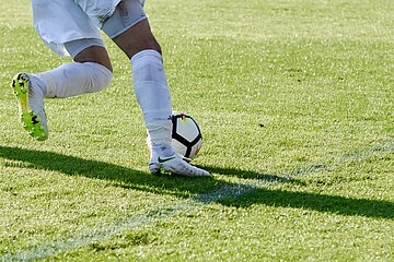 Beine in Sportschuhen spielen einen Fußball auf grünem Rasen, Ball nah am Fuß, Bewegung entlang einer Linienmarkierung.