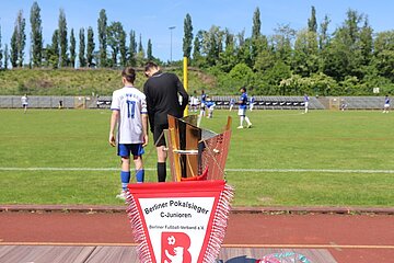Ein Fußballfeld mit Spielern in blauen und weißen Trikots. Im Vordergrund steht ein Pokal mit einem roten Wimpel daran. Auf dem Wimpel steht: "Berliner Pokalsieger C-Junioren Berliner Fußball-Verband e.V." Im Hintergrund sind Bäume und Zuschauer, die das Spiel verfolgen.