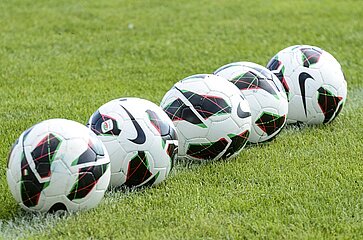 Das Bild zeigt fünf Fußbälle, die nebeneinander auf einer grünen Rasenfläche liegen. Die Bälle sind weiß und besitzen ein modernes Muster aus schwarzen, grünen und roten Elementen sowie ein Nike-Logo. Die Aufnahme ist hell und wirkt freundlich; sie stellt einen typischen Trainings- oder Spielsituation auf einem Fußballfeld dar, ohne Personen oder weitere Gegenstände im Hintergrund.