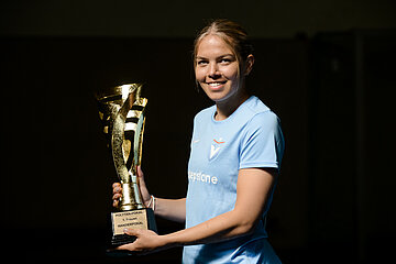 Auf dem Foto ist eine Person zu sehen, die ein hellblaues Trikot trägt. Die Person hält einen goldfarbenen Pokal in den Händen. Der Pokal hat ein modernes, geschwungenes Design und glänzt im Licht. Auf dem Sockel des Pokals ist eine Plakette angebracht. Der Hintergrund des Bildes ist dunkel, wodurch die Person und der Pokal besonders gut sichtbar sind.