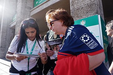 Das Foto zeigt drei Personen, die dicht beieinanderstehen und sich offenbar in ein Gespräch oder eine gemeinsame Aktivität vertieft haben. Die Szene spielt im Freien, vor einem Gebäude mit hellgrauen Steinsäulen und grünen Wegweisern im Hintergrund. Links steht eine Person in einem weißen Shirt mit grünem Akkreditierungsband. Sie hält ein Tablet oder Klemmbrett in beiden Händen und scheint etwas darauf zu zeigen oder zu erklären. Rechts steht eine Person im dunkelblauen Fußballtrikot einer bekannten Vereinsmannschaft. Über ihren Arm hängt ein rotes Kleidungsstück, und sie lehnt sich leicht nach vorn, um besser zu sehen, was gezeigt wird. Zwischen beiden steht eine dritte Person, die ein Heft oder Magazin hochhält. Auf dem Titel ist eine großformatige Überschrift zu lesen und darunter ein Foto mehrerer Menschen. Die Situation wirkt freundlich und kooperativ – als würden die drei Personen Informationen austauschen, etwas überprüfen oder gemeinsam etwas planen, möglicherweise im Kontext eines Fußball-Großereignisses.