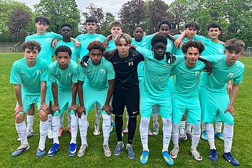 Jugend‑Fußballteam in türkisfarbenen Trikots posiert auf Rasenplatz, vorne gebeugt, hinten stehend, mit Bäumen im Hintergrund.