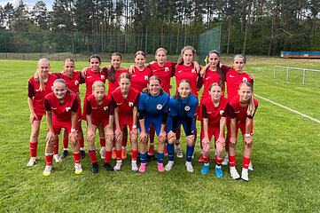 The photo shows a group of 15 people posing in two rows on a soccer pitch. The front row consists of eight people dressed in red jerseys and black shorts. Two people in the middle are wearing blue shirts and shorts. The back row consists of seven people also wearing red jerseys and black shorts. They are all standing or squatting on green grass with a forest in the background.