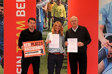 Drei Personen präsentieren ein Antirassismus‑Plakat und Dokumente vor farbigen Sportmotiven. Foto: BFV