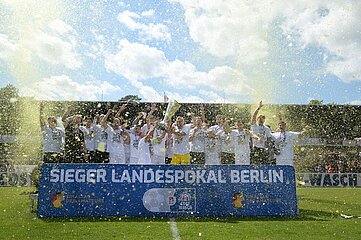 The photo shows a jubilant soccer team celebrating winning the Berlin State Cup. The players are wearing white jerseys and standing behind a striking blue banner with the words "SIEGER LANDESPOKAL BERLIN". Some team members proudly hold up the trophy, while others celebrate the moment with their arms raised. In the background, spectators can be seen in the stands watching the action. The festive and exuberant atmosphere of the moment is further enhanced by sprays of champagne and confetti.