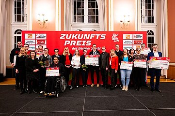 The photo shows a large group of people standing together on a stage in a festive-looking hall. Behind the group is a large red wall with white lettering and logos. The most prominent text reads "Future Prize of Berlin Sports". Several other logos of partners and organizations are visible, including "LSB Berlin", "rbb" and "Berliner Morgenpost". The people stand in two rows next to each other, some holding large symbolic checks, certificates or bouquets of flowers. Among the honorees is also a person in a wheelchair, who stands in the front area and also presents a large cheque. The attire of the people present is predominantly festive or business attire. The room has a prestigious appearance: high, brightly painted walls, large windows with long curtains and classic wall lamps characterize the room. The floor is partially covered with a dark stage platform on which the group stands. Overall, the image conveys the atmosphere of a festive awards ceremony where various people are honored for their achievements or projects in the field of sport.