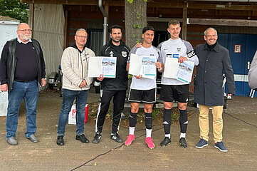 Auf dem Foto sind sechs Personen zu sehen, die nebeneinander stehen. Drei Personen in der Mitte tragen Sportkleidung und halten jeweils eine Urkunde in den Händen – sie scheinen für eine sportliche Leistung ausgezeichnet worden zu sein. Die anderen drei Personen stehen links und rechts neben den Sportlerinnen und tragen Alltagskleidung. Alle Personen stehen aufrecht und blicken in Richtung Kamera. Im Hintergrund ist ein Gebäude mit einer Holzwand zu erkennen, daneben steht ein Baum, der teilweise ins Bild ragt. Die Szene spielt sich im Freien ab, bei Tageslicht.