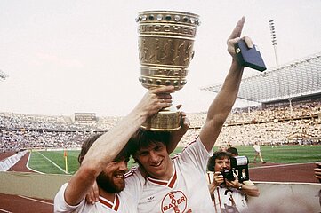Auf dem Foto befinden sich zwei Fußballspieler in weißen Trikots mit roten Details. Sie stehen auf einem großen Fußballfeld in einem voll besetzten Stadion. Einer der Spieler hält einen großen goldenen Pokal mit beiden Händen über seinem Kopf. Der Pokal ist reich verziert und wirkt schwer und massiv. Der andere Spieler hebt den rechten Arm triumphierend in die Höhe und hält ein kleines blaues Etui oder Kästchen in der Hand – möglicherweise eine Medaille. Im Hintergrund sind die Laufbahn und weite Teile der Tribünen sichtbar, die dicht mit Zuschauerinnen und Zuschauern gefüllt sind. Die Stimmung wirkt festlich und jubelnd, als hätten die Spieler gerade ein bedeutendes Finale gewonnen. Rechts am Bildrand steht eine Person mit einer Kamera und filmt die Szene. Die Beleuchtung ist natürlich, und der Himmel ist leicht bewölkt.
