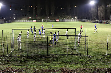 Das Foto zeigt eine nächtliche Fußballszene auf einem beleuchteten Spielfeld. Im Vordergrund ist ein Tor mit Netz zu sehen, um das sich mehrere Spieler gruppiert haben. Einige Spieler tragen weiße Trikots, andere blaue Trikots. Direkt vor dem Tor steht ein Spieler in gelbem Trikot, vermutlich der Torwart. Die Spieler wirken konzentriert, als ob sie sich auf eine Standardsituation wie eine Ecke oder einen Freistoß vorbereiten. Der Rasen ist grün, aber leicht uneben und zeigt Spuren vom Spiel. Im Hintergrund sind weitere Spieler verteilt, ein Schiedsrichter in rotem Trikot ist ebenfalls sichtbar. Hinter dem Spielfeld stehen hohe, kahle Bäume, und am oberen Bildrand leuchten einige Fenster von Gebäuden. Die Szene wird von starken Flutlichtstrahlern erhellt, die deutliche Schatten auf dem Boden werfen.