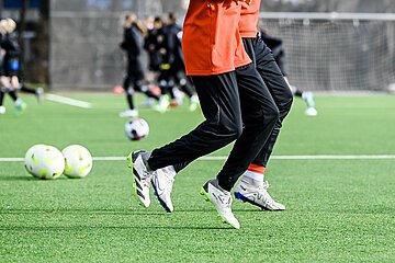 Auf einem Fußballplatz laufen zwei Personen in Sportkleidung seitlich durchs Bild, während im Hintergrund weitere Spielende und mehrere Bälle für eine Trainingseinheit bereitliegen.