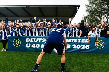Eine größere Gruppe von Personen in blau-weiß gestreiften Fußballtrikots steht auf einem Fußballplatz hinter einer Barriere und hebt die Arme. Im Vordergrund steht eine Person mit dem Rücken zur Kamera in dunkelblauer Sportkleidung mit der Rückennummer 22. Zwischen der Person im Vordergrund und der Gruppe befindet sich eine grüne Werbebande mit dem DFB-Logo und der gut lesbaren Aufschrift: „Deutscher Meister Ü 40 Männer 2026“. Das Foto wurde auf einer Rasenfläche vor einer überdachten Tribüne aufgenommen. Im Hintergrund sind weitere Personen, Bäume und Teile der Sportanlage zu sehen. Die Farben Blau, Weiß und Grün prägen das Bild. Die Szene zeigt einen gemeinsamen Moment im Umfeld eines Fußballwettbewerbs.