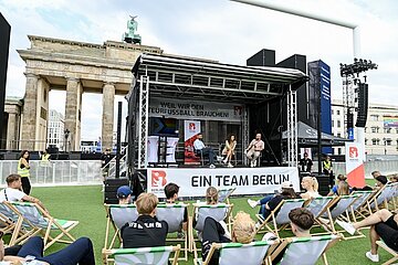 In front of the Brandenburg Gate there is a stage with three people in a round table discussion. The banner on the stage reads "EIN TEAM BERLIN". In front of it, several people are sitting in deckchairs on a green area and watching. Loudspeakers and a large screen can be seen in the background.