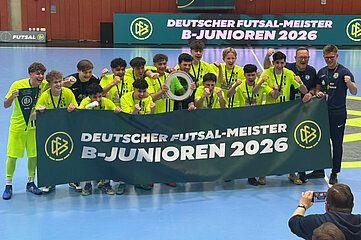 Das Foto zeigt eine Gruppe von Personen auf einem Hallenspielfeld, die eng zusammenstehen und gemeinsam posieren. Alle tragen leuchtend gelbe Sportkleidung, während zwei Personen am rechten Bildrand dunkle Trainingskleidung tragen. Die Gruppe hält ein großes, dunkelgrünes Banner vor sich. Auf dem Banner steht in weißen und gelben Buchstaben: „DEUTSCHER FUTSAL-MEISTER B-JUNIOREN 2026“ Links und rechts auf dem Banner befindet sich das Logo des Deutschen Fußball-Bundes (DFB). In der Mitte der Gruppe hält eine Person einen runden Pokal oder eine Trophäe in die Höhe. Mehrere Personen tragen Medaillen um den Hals. Der Hallenboden ist hellblau, und im Hintergrund hängt ein weiteres großes Banner mit der gleichen Aufschrift wie auf dem Frontbanner. Auf der linken Seite erkennt man zusätzlich ein kleines Logo des Futsal-Wettbewerbs. Im Vordergrund rechts kniet eine Person, die mit einem Handy oder einer Kamera ein Foto oder Video der Gruppe aufnimmt. Die Szene vermittelt den Moment einer Siegerehrung nach einem sportlichen Wettbewerb.