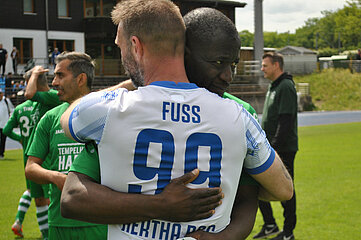 Auf dem Foto sind zwei Fußballspieler zu sehen, die sich freundschaftlich umarmen. Der Spieler im Vordergrund trägt ein weißes Trikot mit blauen Akzenten. Auf seinem Rücken steht der Name „FUSS“ sowie die Rückennummer 99 und der Vereinsname „HERTHA BSC“. Der andere Spieler trägt ein grünes Trikot. Beide Spieler stehen auf einem Fußballfeld. Im Hintergrund sind weitere Personen zu erkennen: Ein Spieler in einem grünen Trikot sowie eine Person in schwarzer Kleidung am Spielfeldrand. Die Szene vermittelt einen Moment des sportlichen Miteinanders und der gegenseitigen Anerkennung.  
