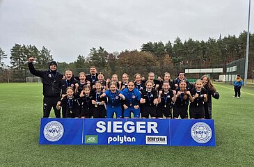 Das Foto zeigt eine Fußballmannschaft auf einem Kunstrasenplatz. Die Gruppe steht dicht beieinander hinter einem großen blauen Schild mit der weißen Aufschrift „SIEGER“. Auf dem Schild sind außerdem Logos des Nordostdeutschen Fußballverbands (NOFV) sowie der Sponsoren „AOK“, „polytan“ und „DERBYSTAR“ zu sehen. Alle Personen tragen schwarze Sportkleidung, einige haben Medaillen um den Hals. In der Mitte hält eine Person einen Pokal. Im Hintergrund sind Bäume mit herbstlichem Laub, ein hoher Zaun und ein Flutlichtmast erkennbar. 