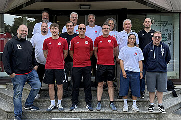 Das Foto zeigt eine Gruppe von insgesamt fünfzehn Personen, die gemeinsam auf einer breiten Außentreppe vor dem Eingang eines Gebäudes stehen. Die Personen sind in zwei Reihen angeordnet: Eine vordere Reihe auf der unteren Stufe und eine hintere Reihe etwas erhöht dahinter. Mehrere Personen tragen einheitliche Sportkleidung. In der vorderen Reihe fallen besonders drei Personen in roten T‑Shirts und dunklen Sporthosen oder -shorts auf. Weitere Personen tragen weiße oder schwarze Oberteile mit sportlichen Logos oder Vereinsabzeichen. Einige Kleidungsstücke zeigen kleine Embleme oder Schriftzüge, die auf eine Organisation oder einen Verein hindeuten. Mehrere Personen tragen Turnschuhe oder lockere Freizeitkleidung wie Jeans, Kapuzenjacken oder Shorts. Die Körperhaltungen sind überwiegend aufrecht und nach vorne zur Kamera gerichtet. Einzelne Personen haben die Arme locker an den Seiten oder vor dem Körper verschränkt, andere halten die Hände in den Taschen. Einige tragen Brillen, und vereinzelt sind kurze Bärte oder unterschiedlicher Haarschnitt sichtbar. Im Hintergrund befindet sich die Fassade eines Gebäudes mit Glasflächen und einer Überdachung. Durch die Fenster sind spiegelnde Oberflächen sowie Teile des Innenraums erkennbar. Auf den Glasflächen sind teilweise Aufkleber oder Schriftzüge angebracht, die jedoch nur teilweise lesbar sind. Die Szene wirkt wie ein Gruppenfoto einer Mannschaft, eines Vereins oder einer gemeinschaftlichen Veranstaltung im sportlichen Kontext.