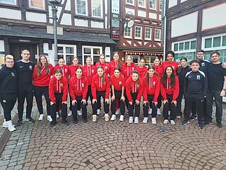 Gruppe in roten Hoodies und dunklen Hosen steht auf einem gepflasterten Platz vor Fachwerkhäusern.