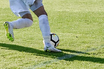 The photo shows a close-up of two legs of a person on a green lawn. The person is wearing light-colored sports shoes with colored accents as well as long, light-colored sports socks and short sports shorts. A soccer ball lies directly in front of one of the feet and is apparently being guided or stopped. The other foot is slightly raised, as if the person is in motion. The grass is evenly mown and sunlight is shining on it, creating clear shadows on the ground. There are no other people or objects to be seen; the image detail focuses exclusively on the lower half of the body, the ball and the movement situation in the sports context.