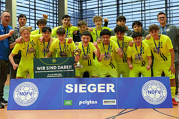 Das Foto zeigt eine große Gruppe von jungen Sportlerinnen und Sportlern, die gemeinsam in einer Sporthalle posieren. Die Gruppe steht dicht beieinander und bildet zwei Reihen. Alle Personen tragen einheitliche, leuchtend gelbe Sportkleidung mit dunklen Streifen und haben Medaillen um den Hals hängen. Vor der Gruppe steht ein breites, blaues Schild mit der Aufschrift „SIEGER“. Auf dem Schild befinden sich außerdem mehrere Logos, darunter das Logo des Nordostdeutschen Fußballverbands (NOFV) sowie Sponsorenlogos. In der Mitte der vorderen Reihe hält eine Person eine Trophäe in der Hand. Direkt daneben wird ein großes, grünes Schild hochgehalten. Links und rechts neben der Gruppe stehen zwei Menschen in blauer oder grauer Sportkleidung, die ebenfalls mitjubeln. Der Hintergrund zeigt eine helle Sporthalle mit hohen Fenstern und dunklen Wandbereichen. Der Boden besteht aus typischem Hallenbelag mit Linien für verschiedene Sportarten. Die gesamte Gruppe zeigt eine feierliche Siegerpose, teilweise mit erhobenen Armen oder geballten Fäusten.