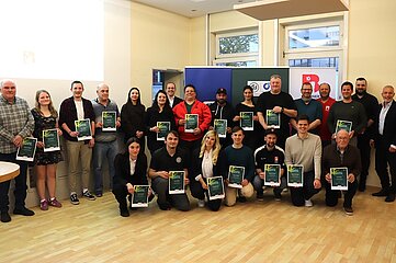 Das Foto zeigt eine Gruppe von etwa 30 erwachsenen Personen, die in zwei Reihen in einem hellen Innenraum stehen und knien. Der Raum wirkt wie ein Veranstaltungs‑ oder Seminarraum mit hellem Holzboden, weißen Wänden und großen Fenstern im Hintergrund. Tageslicht fällt von der rechten Seite in den Raum. Im Mittelpunkt stehen die Personen, die fast alle eine gerahmte Urkunde oder ein Zertifikat mit dunklem Hintergrund und grünen Elementen in den Händen halten. Die Tafeln sind überwiegend im Hochformat. Die Personen in der vorderen Reihe knien oder hocken, während die Personen in der hinteren Reihe stehen. Alle blicken in Richtung Kamera. Die Kleidung ist unterschiedlich: Sie reicht von sportlich über casual bis hin zu schlichter Business‑Kleidung. Zu sehen sind verschiedene Oberteile wie Pullover, Kapuzenjacken, Hemden, T‑Shirts und Jacken in Farben wie Schwarz, Grau, Blau, Grün, Rot und Beige. Im Hintergrund stehen mehrere Roll‑ups oder Banner mit Logos und Symbolen in Blau, Grün, Weiß und Rot. Der Text auf den Bannern ist teilweise sichtbar, aber nicht vollständig lesbar. Die Banner deuten auf eine organisierte Veranstaltung oder eine Ehrung hin. Der Gesamteindruck des Bildes ist eine formelle Gruppenaufnahme im Rahmen einer Auszeichnung oder Anerkennung, bei der viele Teilnehmende gemeinsam präsentiert werden.