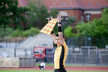 A person serving as a linesman on a sports field raises a flag to signal; in the background, a track, a fence, and buildings are visible, along with plenty of greenery. Photo: Sandra Ritschel