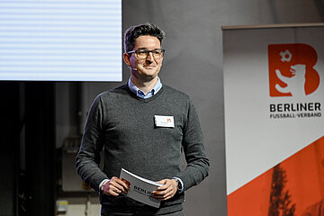 Person steht auf einer Bühne mit Unterlagen in der Hand; rechts daneben ein Roll‑Up mit dem Logo des Berliner Fußball‑Verbands. Foto: Sandra Ritschel