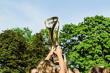 Das Bild zeigt eine Gruppe von Menschen, die gemeinsam einen goldfarbenen Pokal in die Höhe halten. Der Pokal hat ein aufwendig gestaltetes Design. Die Szene spielt im Freien – im Hintergrund sind grüne Bäume und ein klarer blauer Himmel zu sehen. Es ist ein Moment des Feierns nach Siegerehrung. Foto: Sandra Ritschel