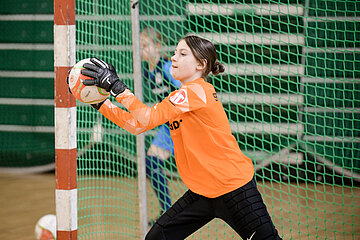 Auf dem Foto ist eine Person zu sehen, die in einer Sporthalle als Torhüter*in beim Hallenfußball agiert. Die Person trägt ein auffälliges orangefarbenes Torwarttrikot, schwarze gepolsterte Torwarthosen und schwarze Torwarthandschuhe. Sie steht direkt vor einem Hallenfußballtor und hält mit beiden Händen einen Ball fest, als würde sie gerade einen Schuss abwehren. Im Hintergrund befindet sich ein weiteres Kind oder eine weitere Person, die in blauer Sportkleidung zu erkennen ist. Das Tor ist mit einem grünen Netz bespannt, und die Umgebung wirkt wie ein typischer Hallensportbereich mit Holzfußboden und Sportbande. Die Szene vermittelt Konzentration, Bewegung und sportlichen Einsatz.