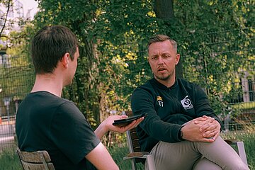 Zwei Personen sitzen draußen im Gespräch, eine Person hält ein Smartphone in der Hand und richtet eine Frage an ihr Gegenüber. Im Hintergrund sind Bäume, Zaun und eine ruhige, grüne Umgebung zu sehen.