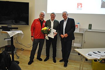 Drei erwachsene Personen stehen nebeneinander in einem hellen Seminarraum. Die Person in der Mitte hält einen Blumenstrauß und eine Urkunde in den Händen. Links steht eine Person in einer roten Sportjacke, rechts eine Person in dunkler Kleidung. Im Hintergrund sind eine Präsentationsfläche mit einem Logo sowie ein großer Bildschirm zu sehen. Neben den Personen stehen Tische mit Unterlagen und Technik.