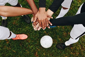Das Foto zeigt eine Gruppe von fünf Personen, die im Kreis stehen und ihre Hände in der Mitte übereinandergelegt haben – ein Symbol für Teamgeist und Zusammenhalt. Alle Personen tragen Sportkleidung, vermutlich Fußballtrikots und -hosen. Die Hände unterscheiden sich in Hautfarbe, was auf eine vielfältige, inklusive Gruppe hinweist. Im Hintergrund liegt ein Fußball auf einem grünen Rasen, was die sportliche Atmosphäre unterstreicht. Die Perspektive des Bildes ist von oben aufgenommen, sodass der Fokus auf den Händen und dem Gemeinschaftsgefühl liegt.