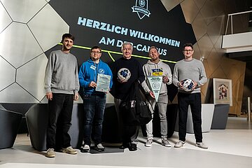In einem modern gestalteten Innenraum stehen fünf Personen nebeneinander vor einer großen digitalen Leinwand mit dem Schriftzug „HERZLICH WILLKOMMEN AM DFB‑CAMPUS“ und einem Verbandslogo; sie halten Urkunden, Fußbälle und eine Sporttasche in den Händen, während im Hintergrund geometrische Wandstrukturen, graue Sitzmöbel und ein gerahmtes Trikot zu sehen sind.