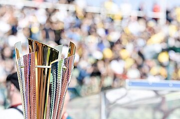 Im Vordergrund des Fotos steht eine Pokaltrophäe aus Metall. Die Trophäe ist hoch und schlank geformt und besteht aus mehreren leicht gebogenen Elementen, die nach oben hin geöffnet sind. Die Oberfläche wirkt glänzend und reflektierend, mit gold- und silberfarbenen Tönen, die je nach Lichteinfall schimmern. Das Material scheint strukturiert zu sein und wirft kleine Lichtreflexe. Im Hintergrund ist unscharf eine große Menschenmenge zu erkennen, vermutlich auf einer Tribüne oder in einem Stadion. Die vielen Farben – unter anderem Gelb, Grün und Weiß – deuten auf Kleidung, Fahnen oder Sitzplätze hin. Einzelne Personen sind nicht erkennbar, da der Fokus des Bildes klar auf der Trophäe liegt. Die Szene findet offenbar im Freien statt, bei Tageslicht. Das Bild vermittelt den Eindruck eines sportlichen Wettbewerbs oder einer Siegerehrung, bei der die Trophäe stellvertretend für Erfolg, Leistung oder einen gewonnenen Titel steht.