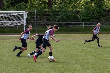 Auf dem Bild ist eine Szene eines Fußballspiels auf einem grünen Rasenplatz zu sehen. Im Vordergrund befinden sich drei Spieler. Alle tragen weiße Trikots mit schwarzen Hosen und lila Ärmeln. Einer der Spieler führt den Ball am Fuß, während die beiden anderen versuchen, ihn zu stoppen oder den Ball zu erobern. Im Hintergrund steht ein Fußballtor mit einem weißen Netz. Dahinter sind eine dichte grüne Hecke und mehrere Bäume zu erkennen, die den Platz begrenzen.