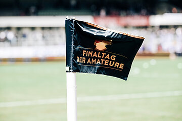A corner flag with the inscription "Final day of the amateurs" stands on the edge of the pitch, the stadium can be seen blurred in the background. Photo: Sandra Ritschel