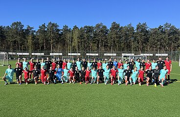 Group photo of several youth football teams on a green artificial turf pitch. Around 40 people stand and kneel in a row, wearing different jerseys in red, turquoise, black and blue. Soccer goals, a fence, parked cars and a dense forest under a blue sky can be seen in the background.