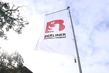 Weiße Fahne mit dem Logo und Schriftzug des Berliner Fußball-Verbands. Das Logo zeigt ein rotes ‚B‘ mit einer weißen Figur, die einen Fußball hält. Die Fahne weht an einem Mast, im Hintergrund sind Bäume, Sonnenlicht und ein teils bewölkter Himmel zu sehen. Foto: BFV