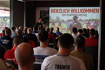 The photo shows a large event hall during a presentation. In the foreground, several rows of chairs are visible, with numerous participants seated on them and looking forward. Most people are wearing athletic clothing, often with club names or labels such as “COACH” on the back. Everyone’s gaze is directed toward the presentation area. In the center, a person stands to the side of the screen and speaks into a handheld microphone. The person is positioned slightly to the left of the projection screen and is clearly visible to the group of attendees. A large projection screen dominates the background. A slide with the heading “WELCOME to the Training Day” is visible on it. Below this heading is an image of a group of people on a soccer field, with a person in the foreground wearing a jersey with “1. FC UNION BERLIN” on the back. Additionally, logos and graphic elements related to soccer are visible. The room is designed to be rather dark, with dimmed walls and ceilings, which makes the bright projection stand out particularly well. On the left side of the image, windows are visible through which daylight streams in. The combination of subdued interior lighting and the strong, colorful projection draws attention to the presentation. Overall, the image conveys a formal training session or workshop in a soccer context, where a large group of participants is listening to a welcome presentation.