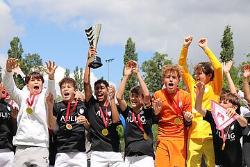 Eine Gruppe von Kindern feiert gemeinsam den Gewinn des Nike Youth Cups. Sie tragen Sportkleidung in verschiedenen Farben – darunter schwarze Trikots mit der Aufschrift „MULIG BAU“, sowie weiße und gelbe Outfits. Viele der Kinder haben Medaillen um den Hals. Der Pokal wird von mehreren Kindern in die Höhe gehalten. Die Gesichter der Kinder sind nicht erkennbar. Im Hintergrund sind Bäume und ein teilweise bewölkter Himmel zu sehen.
