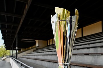 Das Foto zeigt einen modernen Pokal, der in einem leeren Stadion steht. Der Pokal besteht aus glänzendem Metall mit goldenen und silbernen Elementen. Er hat eine elegante, nach oben geöffnete Form. Im Hintergrund sind mehrere Ebenen leerer Tribünen zu sehen, die aus grauen Sitzreihen bestehen. Darüber wölbt sich ein dunkles Stadiondach. Es sind keine Menschen im Bild zu sehen, und die Atmosphäre wirkt ruhig und still – als würde das Stadion auf eine kommende Veranstaltung warten.
